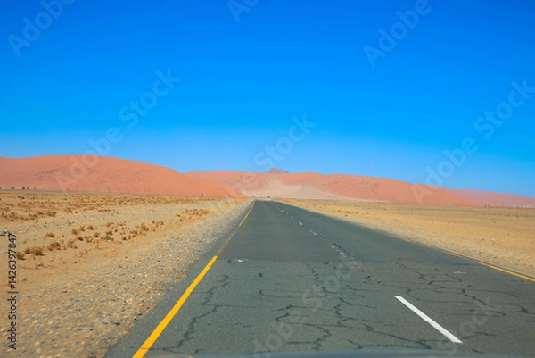 Fototapeta The asphalt road towards Sossusvlei cuts through the rugged and desolate Namibian desert landscape against blue sky. The high red dunes of the Namib Desert rising in the distance. Namibia, Africa.