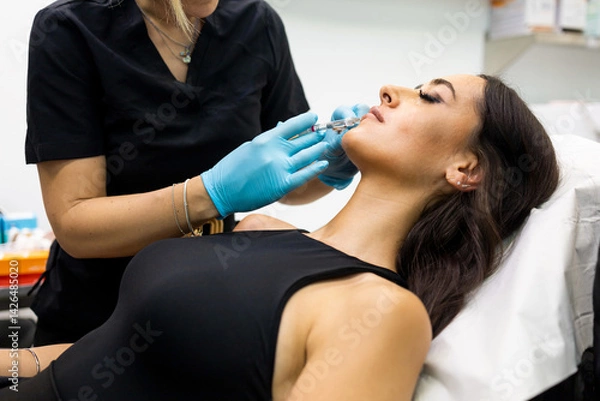Fototapeta An unrecognizable aesthetic doctor injects hyaluronic acid into a girl's lips in a modern clinic. Concept of hyaluronic acid on lips, aesthetic operations on lips.