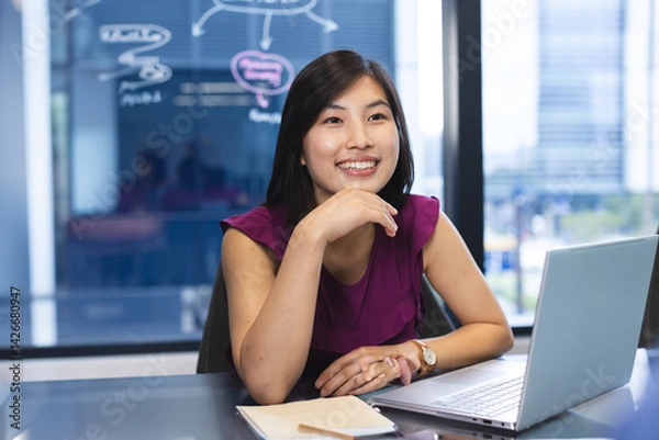 Obraz Asian woman working on laptop at glass desk in office, showing marker diagrams and city view