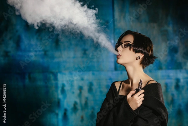 Fototapeta A girl with glasses vaping and releases a cloud of vapor. Model in a black vaper smoke vaporizer on a turquoise background