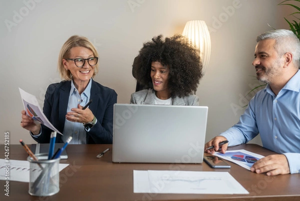 Fototapeta Three busy happy middle aged professional business man and two women executive leaders team using laptop working on computer at work desk having conversation on financial project at meeting in office.