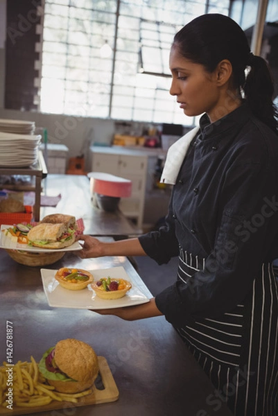 Obraz Indian woman chef plating tartlets and sandwiches on white plate in restaurant kitchen, copy space