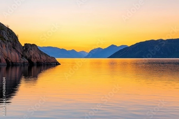 Fototapeta A serene sunset over calm waters, reflecting vibrant orange and yellow hues, surrounded by rugged mountains and cliffs.