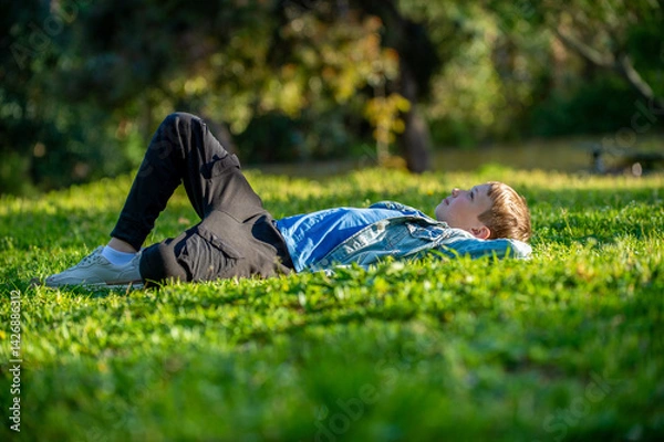 Fototapeta Young cheerful child relaxing in a park. Smiling child laying down on the grass in a garden and dreaming, on a sunny summer day. Happy young boy imagining his future on a warm summer day.
