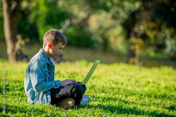 Fototapeta Student with a laptop on his feet is sitting on a green lawn in a park. Young boy with his hands on a computer keyboard in the park in the summer. Thinking student, typing on a keyboard outdoors