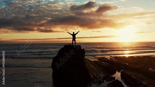 Fototapeta Human with raised hands looks to the sun over horizon in morning while sunrise.  Man stands on a rock against the ocean  beautiful summer sunset. Beauty of nature.  Silhouette of a man against the sky