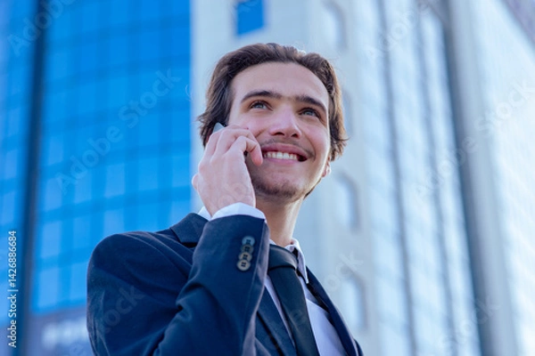 Fototapeta Smiling businessman near a business center calling by cell phone. A happy young businessman talking with a smartphone. Young cheerful businessman near a modern tall glass building with a phone. .