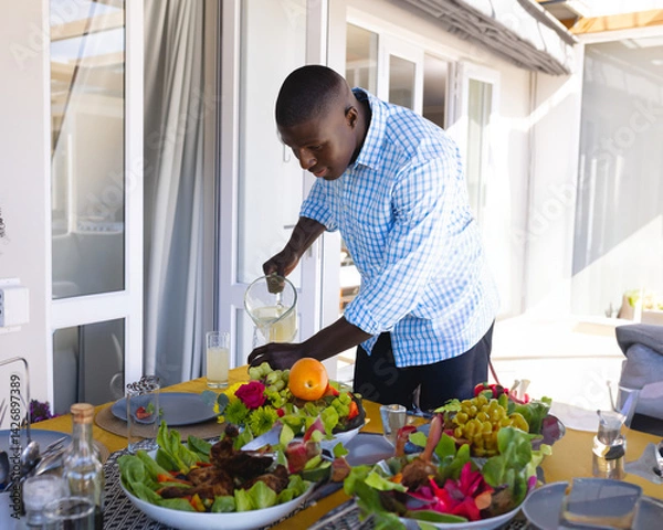 Obraz African American man pouring juice from glass pitcher into glass on patio table, with salad, fruit