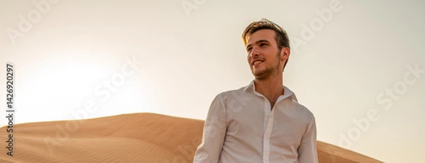 Fototapeta Handsome young man in a white shirt looks into the distance against the background of sand dunes. Portrait of a happy man in nature in the desert.