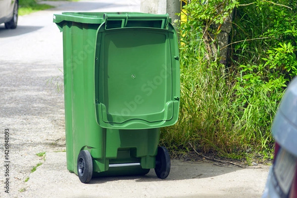 Fototapeta Individual garbage bin for unseparated household waste. A medium-sized green trash container on wheels stands with the lid open, left on the street by workers of the municipal garbage removal service.