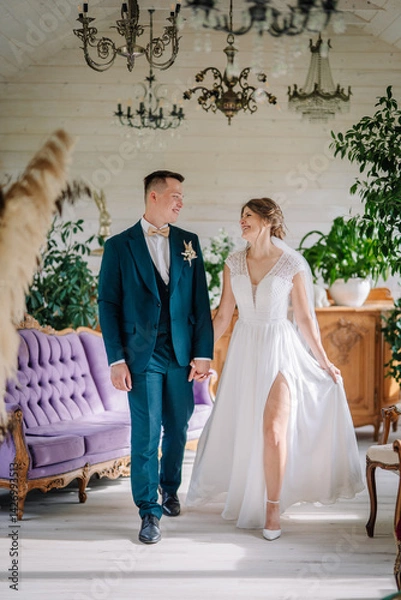 Obraz Bride and groom walk hand in hand, smiling joyfully in a bright, elegant room filled with greenery and vintage decor on their wedding day..