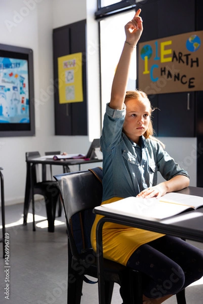 Fototapeta Girl child raising hand in modern classroom, holding pen over open notebook on desk