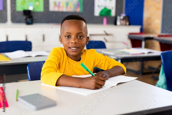 Fototapeta African American child writing in open notebook at school desk, using green pen and smiling