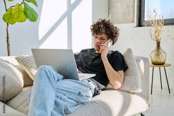 Fototapeta Handsome young man with laptop using smartphone sitting on sofa at home