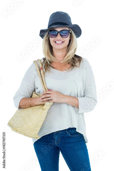 Fototapeta Middle-aged woman standing and smiling in indoor studio setting, with sun hat and straw tote bag