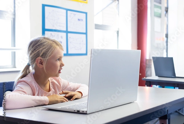 Fototapeta girl child working on silver laptop at desk in modern classroom, with math posters and daylight