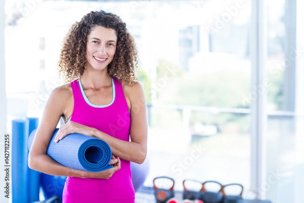 Obraz Woman holding rolled-up yoga mat and smiling in fitness studio, with exercise ball and kettlebells
