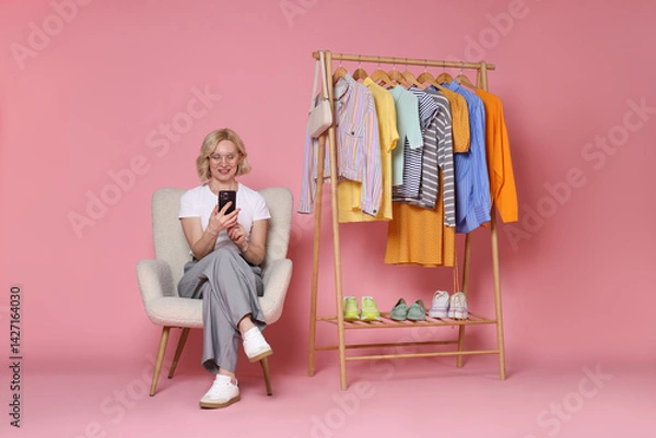 Fototapeta Smiling fashion stylist with smartphone and clothing rack on pink background