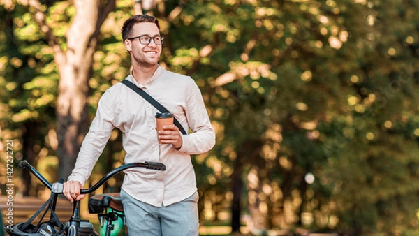 Fototapeta Modern lifestyle and ecological transport. Happy hipster in glasses holding coffee and walking with bike in park, copy space