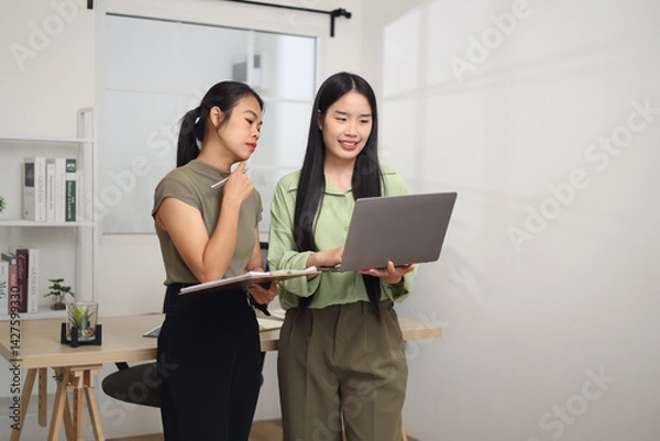 Obraz Portrait of Two Asian business woman standing in an office, holding documents and discussing work. Business ideas and strategies
