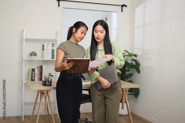 Obraz Portrait of Two Asian business woman standing in an office, holding documents and discussing work. Business ideas and strategies