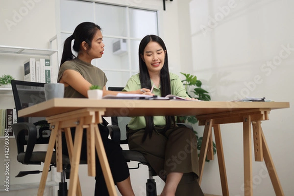 Obraz Low angle view shot, Two female colleagues are sitting together at desk, reviewing documents and discussing work in the office.