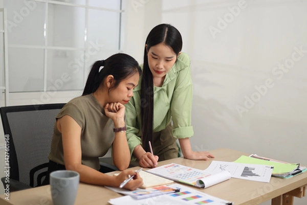 Fototapeta Portrait of Two female colleagues are working together in an office. One stands explaining the work, while the other sits and listens with interest.