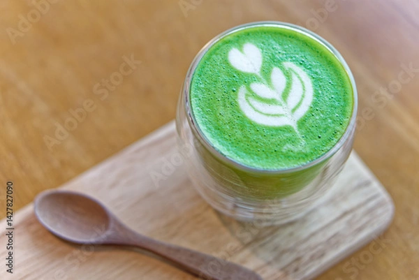 Fototapeta Matcha latte with heart-shaped latte art in clear glass on wooden tray. Hot matcha latte with delicate heart and leaf latte art served in double-wall glass cup on wooden tray with a spoon on the side