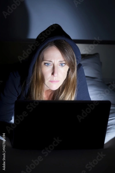 Obraz Close-up of young woman in hoodie under dim light, working on laptop, notifications flashing on phone
