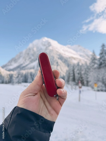 Fototapeta Hand holding a red multi-tool knife with snowy mountain peak and forest in the background