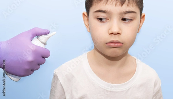 Obraz kid using drops or saline spray.child looking confused and weird at bottle with physiological seawater.hand in surgical glove holding spray against blue backdrop.doctor heel boy from stuffy nose.
