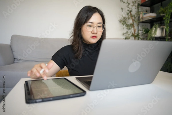 Obraz Asian woman sits on the floor typing laptop and using digitalized pen writing on tablet in living room