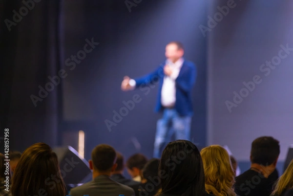Obraz Presenter speaking on stage with blurry focus, addressing audience in a conference setting.