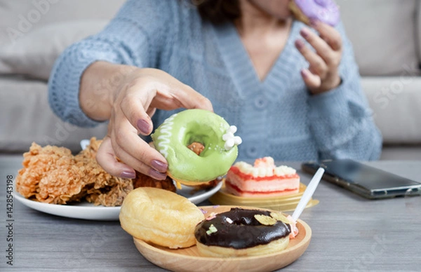 Obraz woman’s hand reaches for a donut on table that full of junk food like fried chicken, cake, sweet drinks, and desserts, showing signs of sugar addiction, eating too much, and eating unhealthy food 