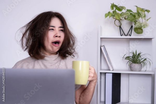 Obraz Tired Asian woman sitting at her office desk, yawning and holding a cup of coffee, feeling sleepy and low energy in the morning due to a sleepless night, hangover, or trouble falling asleep