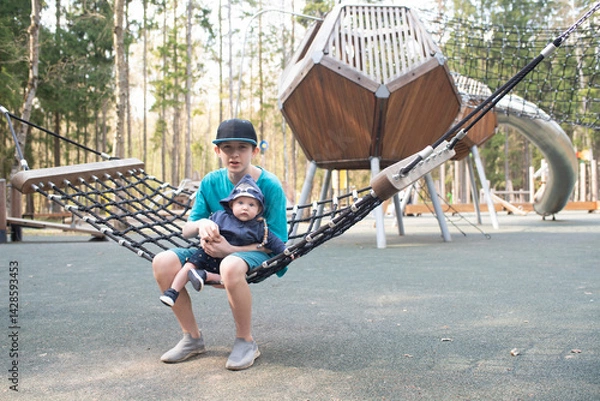 Fototapeta A 12 year old boy with his baby brother on his knees swings on a hammock swing at a playground in Borovsky Park in Moscow.
