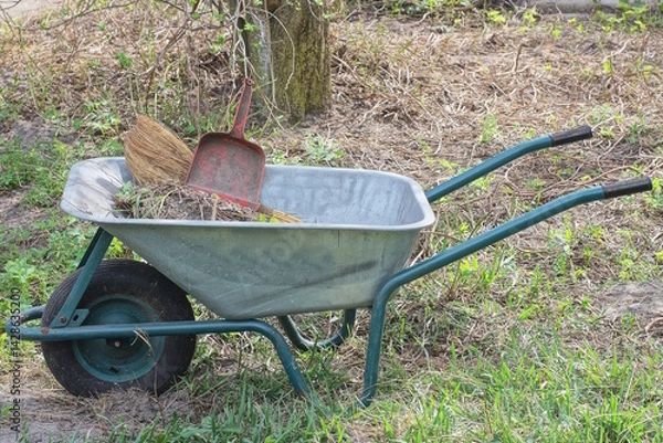 Obraz one large metal garden wheelbarrow with a dustpan and a red dustpan stands on the ground and green grass in a spring garden