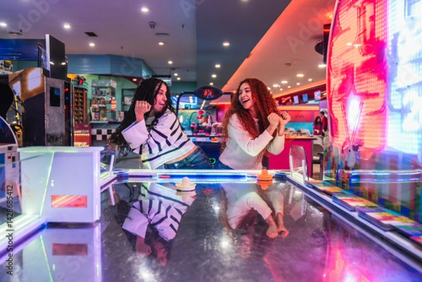 Fototapeta Two happy friends are enjoying their time playing air hockey in a vibrant arcade videogame room, celebrating their friendship and the thrill of the game