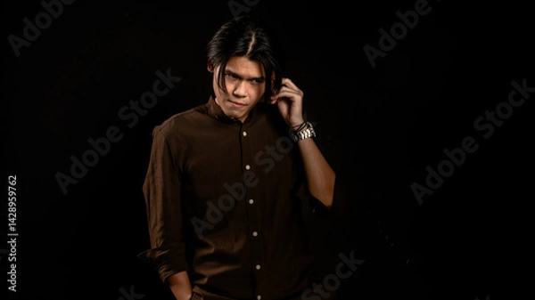 Fototapeta Asian man in dark brown button-up shirt standing in studio with hand touching ear and intense gaze, posing confidently against black background, ideal for fashion, branding, or edgy lifestyle content