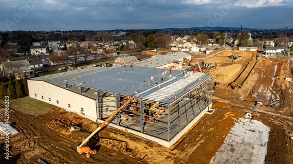 Fototapeta Work continues on a large commercial structure, with workers actively installing roofing materials and securing the steel framework under a cloudy sky. Surrounding fields and homes are visible.