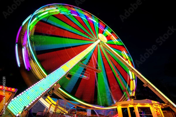 Obraz Colorfully illuminated Ferris wheel at a funfair in Germany at night. Atmospheric long exposure with motion blur. Light traces of the rotating carousel from extreme wide angle frog's perspective. 
