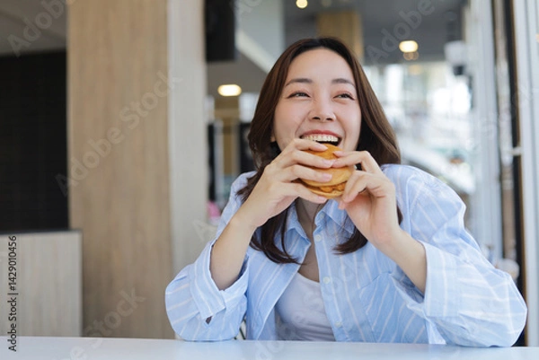 Obraz A cheerful young Asian woman is smiling brightly while holding a burger with both hands, ready to take a bite. She is enjoying her meal at a modern cafe or restaurant.