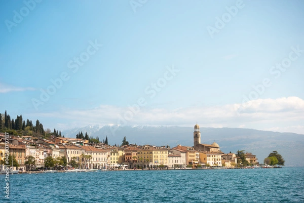 Fototapeta Panoramic view of coastal town Salò by Lago di Garda lake with iconic church tower in front of alps mountains in background on clear day