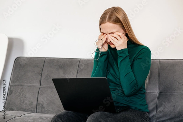 Fototapeta 
Young woman sitting at desk, rubbing tired eyes from screen fatigue and stress. Concept of digital eye strain, overwork, computer vision syndrome, burnout, and remote work pressure 
