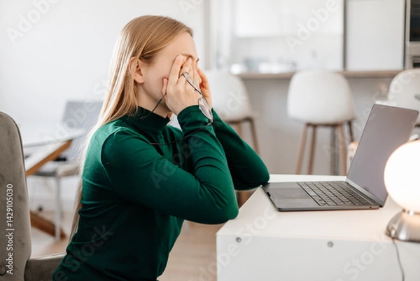 Fototapeta 
Young woman sitting at desk, rubbing tired eyes from screen fatigue and stress. Concept of digital eye strain, overwork, computer vision syndrome, burnout, and remote work pressure 
