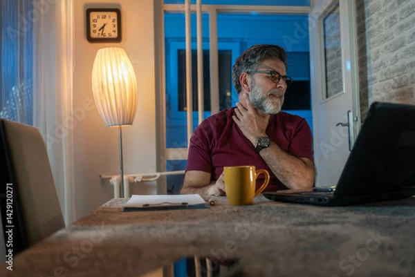 Fototapeta Sick mature man at home office in evening, touching throat in pain while using laptop. Flu, cold, virus, work from home, health issue and stress concept.