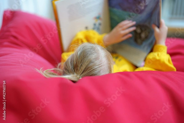 Fototapeta Child reading book while relaxing on soft red bean bag in comfortable indoor space during cozy afternoon