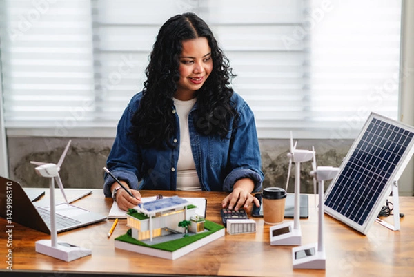 Obraz Female architect analyzes energy efficiency by calculating solar panel output, wind turbine capacity, and energy demand using home models, promoting renewable energy use in smart home design.