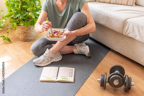 Fototapeta Sitting on a fitness mat, a mature woman enjoys a healthy meal after her home workout, her meal plan and dumbbells resting nearby, symbolizing her commitment to a balanced lifestyle