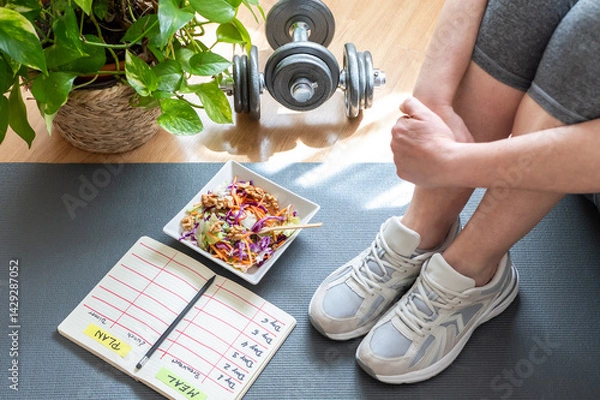 Fototapeta Sitting on a yoga mat, a woman wearing sneakers reviews her meal plan and workout schedule, accompanied by a healthy salad and dumbbells, promoting a balanced lifestyle